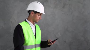 Telefonlu Arap mühendis. Gri arka planda akıllı telefonu olan bir inşaatçı. İnşaatçının ya da mimarın kasketi ve inşaat üniformaları. İnşaat satış konsepti. Koruyucu üniforma satışı