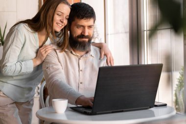 Evde dizüstü bilgisayarı olan mutlu genç bir çift. Evde ya da otel odasında arka planda penceresi olan bir masa.