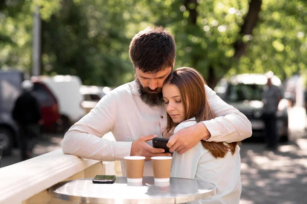 Couple in love drinking coffee in a street cafe in summer, man hugging woman and using a smartphone to call or search the internet, copy space