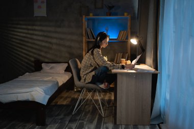 Asyalı genç iş kadını ya da öğrenci. Geceleri çizelge ve dokümanlara sahip, fazla mesai yapan, kulaklık takan bir öğrenci. Akşamları iş, eğitim ve teknoloji kavramı.