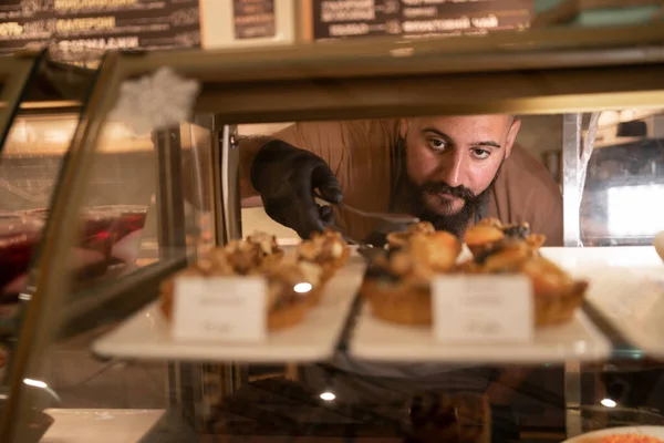 Indian man choosing variation of delicious cake in bakery showcase fridge at cafe. Male customer buying sweet cake at coffee shop. Small business restaurant food concept