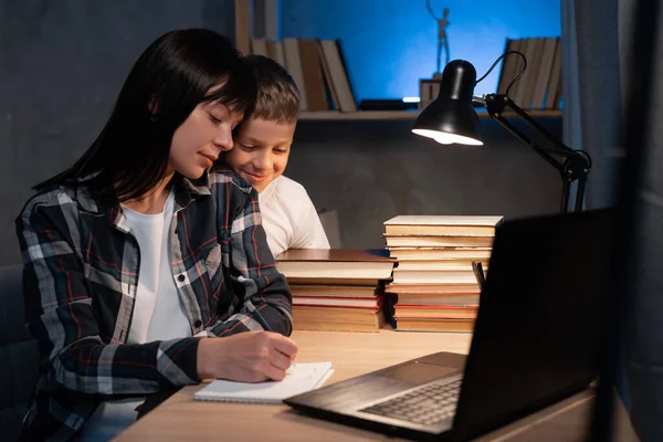 Busy mom works at home with son at night. Mother is watching on laptop while working remotely and writing notes at the table.