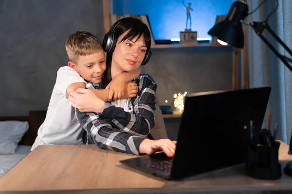 Working at home with kids. Young focused woman mother using laptop and thinking about work task working while boy son gently hugs her. Mum and son at night at room near desk. Childcare concept