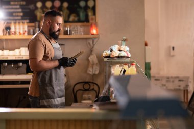 Dijital tablet kullanarak ve ekrana bakarak başarılı bir küçük işletme sahibi. KOBİ konsepti sayarken vitrinin önünde duran sakallı ciddi bir barista.