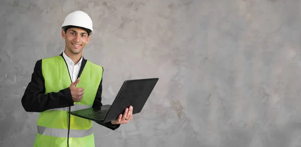 Arabic civil engineer using laptop. Engineering and construction concept. Happy engineer with laptop computer. Man in hard hat. Architect in yellow vest on grey background. Banner, space for text