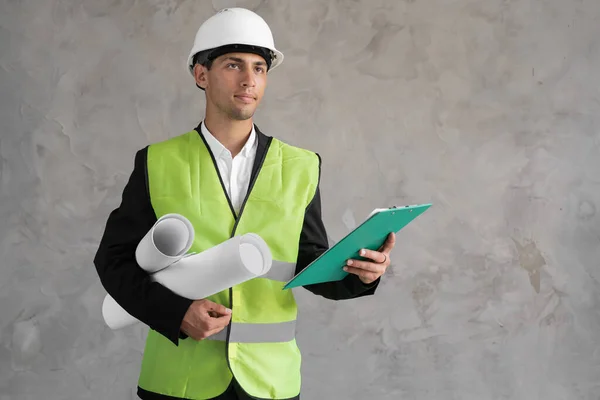 Confident young arabic male construction engineer in hardhat and waistcoat holding rolled blueprint and looking at camera thinks against grey background, copy space