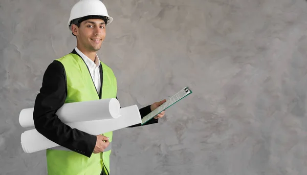 Young arabic civil engineer holding paperwork blueprint roll standing post in studio. Content male construction architect in suit and hardhat with rolled blueprints on isolated grey background. banner