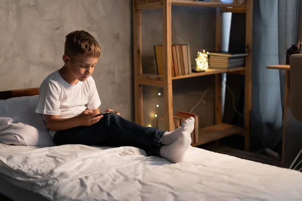 Boy child sitting on bed in the evening watching cartoons or playing mobile games on phone during leisure time. Gadget addiction among children