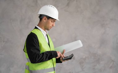 Yeni bir evin maliyeti. Gri arka planda çizimleri ve hesap makinesi olan Müslüman genç bir adam. İnşaat tahmini. İnşaat alanında beyaz kasklı bir Mimar..