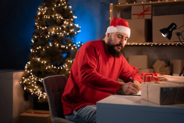 Genç acemi sakallı Hintli girişimci, Noel Baba kostümlü küçük işletme sahibi gece evde çalışmaktan yorulmuş, paket teslimat adresini kalemle dolduruyor. Noel indirimi. Boşluğu kopyala.