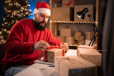 Young beginner bearded hindu man entrepreneur, small business owner in santa claus costume tired working at night in home office, tying red bow on delivery box. christmas sale. copy space.