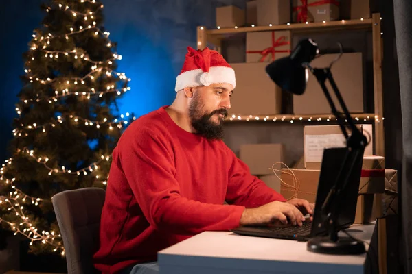 small business owner Indian freelance Santa working in home office using computer, online marketing packaging box and gifts for Christmas delivery, SME e-commerce, Christmas sale