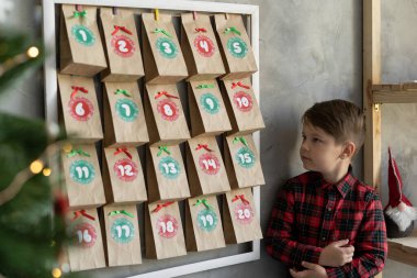 Duvarda asılı duran el yapımı takvim, şekerli el işi çantaların yanında duran çocuk, Noel bebek sevinci, kış gelenekleri