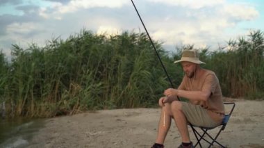 a man fisherman at sunset in a hat on the lake catches fish with a fishing rod against the background of reeds. sits on a chair stringing bait on a hook. Fishing holiday concept. copy space