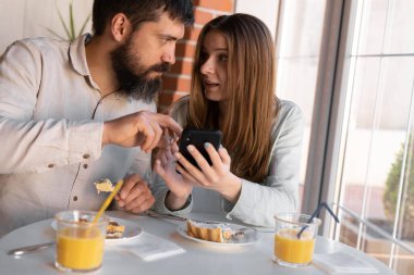 Portrait of happy cute young couple looking at phone and smiling while eating and sitting in a cafe. Joyful couple using cellphones and laughing while having breakfast in cozy kitchen at home