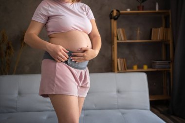 Pregnant woman in rose pajamas with orthopedic support belt, pregnancy bandage, on gray background.