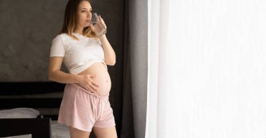 Young pregnant woman drinking water, future lady relaxing at home with glass, copy space. Pregnancy, healthcare, thirst concept. banner, empty space