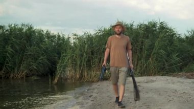 caucasian bearded man catches fish in the evening at sunset on a summer day on a river in the countryside, walks along the shore, holds a landing net and a fishing rod in his hands. copy space.