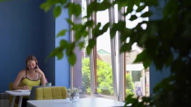 A young female freelancer sits in a cafe, talking on a mobile phone and writing diary entries. Day planning concept. copy space.