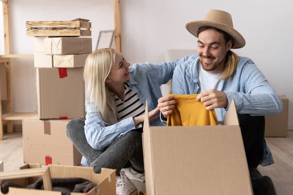 a young cheerful couple in a new apartment on the day of the move sit on the floor unpack things the woman puts on a hat to the man. moving to a new apartment. Lots of packing boxes around them.