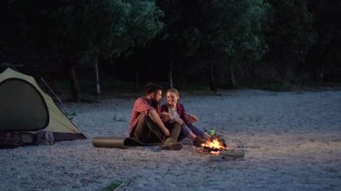 Happy tourist couple camping in the wild eating hot dogs and kissing. Couple cuddling by the campfire and roasting a delicious snack near their tent. Cheerful young woman and man by the fire