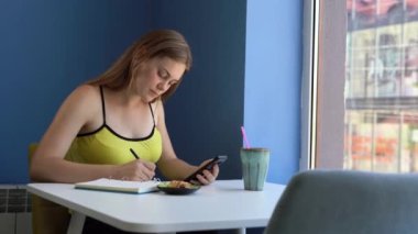 A young caucasian woman sits in a yellow t-shirt in a cafe with blue walls and writes with a pen in a diary, holds a smartphone in her hands. Day planning concept