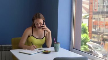 A young female freelancer sits in a cafe, talking on a mobile phone and writing diary entries. Day planning concept. copy space.