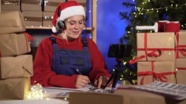 Noel Baba gibi giyinmiş beyaz bir kadın internetten mal teslimatı için kulaklık ve akıllı telefon kullanıyor. Büyük yeni yıl konsepti ya da Noel malları satışı..