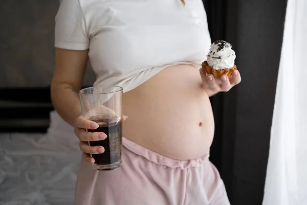 close up image of pregnant woman with cake and sparkling cola standing at home. Pregnancy and unhealthy eating concept
