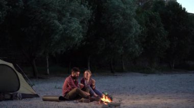 Night summer camping on the seashore. Two young tourists a man and a woman are sitting and eating a hot dog near the fire near the tent. Tourism, friendship and beauty of nature concept.