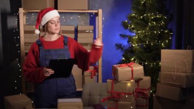 Küçük işletme bırakma konsepti. Kafkasyalı kadın çevrimiçi dükkan sahibi gece evde Noel Baba gibi giyinmiş teslimat için kutuları sayıyor ve işaretliyor. Büyük Noel İndirimi.