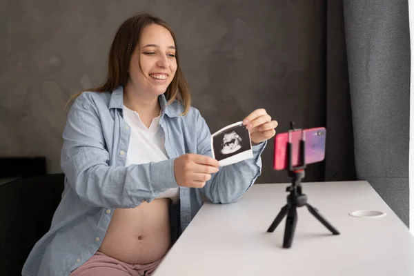 pregnant woman blogger showing ultrasound picture at camera, sitting at home, young future mom demonstrating scan on smart phone, waiting for first baby, pregnancy and motherhood concept