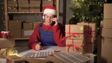 small business worker selling goods in santa costume and overalls table near packed boxes with red bows takes an order by phone makes notes on the form. Big Christmas Sale. Dropshipping and delivery