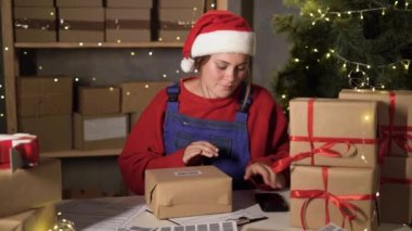 A small business worker in a santa claus costume sticks a barcode on a box with a red bow and scans it with a smartphone. Big Christmas Sale. Dropshipping and delivery of goods