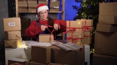 Caucasian woman dressed as santa claus businessman working in office in evening. Packing and delivery service for goods during Christmas sale, light bulbs glow, tie a bow on the box. Small business