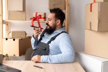 Millennial Indian man business worker using smartphone at storehouse, records message using voice assistant on mobile phone. E-commerce male business owner using phone in store warehouse.