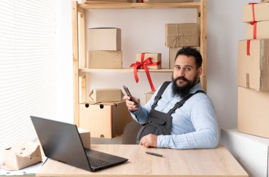 Indian male small business owner using mobile app on smartphone checking parcel box. Warehouse worker, seller holding phone do retail drop shipping package postal parcel. SME, shopping online concept