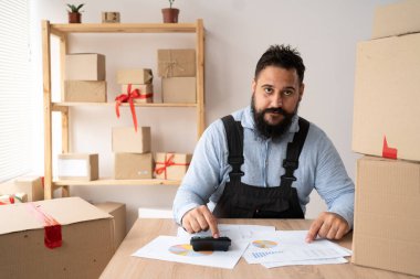 Portrait of Indian man, e-commerce employee sitting in the office full of boxes on the table using a calculator, SME business, e-commerce, technology and delivery business.