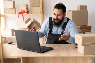 Evil bearded male business owner sitting at table and calling while browsing online on laptop. sme business freelance working in home office using computer, online marketing packaging box delivery,