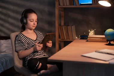 Caucasian schoolgirl watching something on tablet computer at night, preteen girl in wireless headphones using digital tablet at home for studying, video call to teacher or parent, talking at webcam