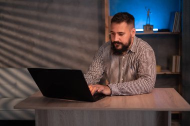 Young Indian ethnic businessman developer or web designer typing on laptop distance working online late at night at home or in dark office, using laptop sitting at workplace desk.