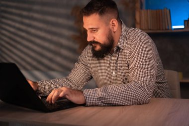 Young Indian ethnic businessman developer or web designer typing on laptop distance working online late at night at home or in dark office, using computer sitting at workplace desk.