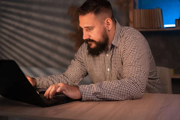 Young Indian ethnic businessman developer or web designer typing on laptop distance working online late at night at home or in dark office, using computer sitting at workplace desk.