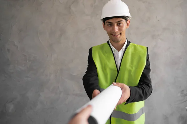 Young arabic architect man with helmet and holding blueprints isolated on grey background, construction engineer in a hard hat and an vest. Builder with drawings.