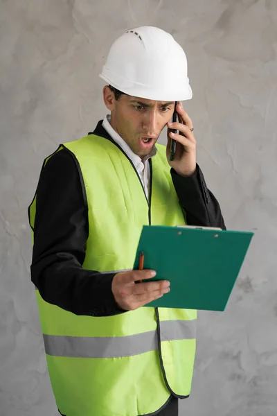 Angry Arabic architect screaming at phone during call making angry gesture with hand isolated on grey studio background. Despair engineer with smartphone. Worker with helmet and clip board.