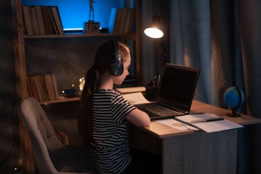 little girl in wireless headset using laptop, studying online at home at night, interested happy student typing on keyboard looking at pc screen, watching webinar, online course, doing homework
