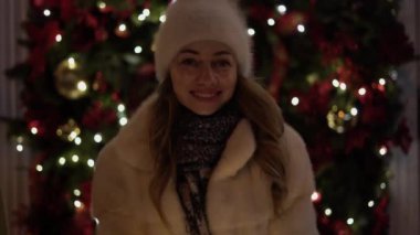 A beautiful young woman looks into the camera smiling and holds glowing garlands in her hands. Festive Christmas market on the background. Model looks at the camera, wears a knitted cap