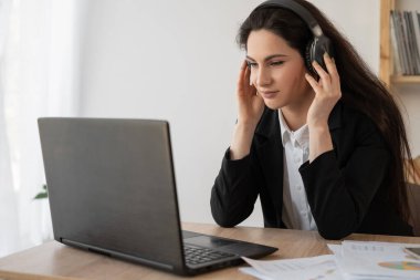 Serious businesswoman in headphones watching webinar on laptop, learning studying computer course, making call, participating online conference, copy space