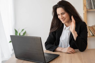Image of a beautiful brunette woman posing sitting indoors office using laptop computer talking waving. Web camera distant conversation using computer app, enjoying video call online meeting in office
