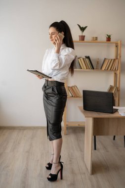 Elegant asian businesswoman using mobile phone after working on digital tablet, standing in office interior, woman manager talking to client using smartphone